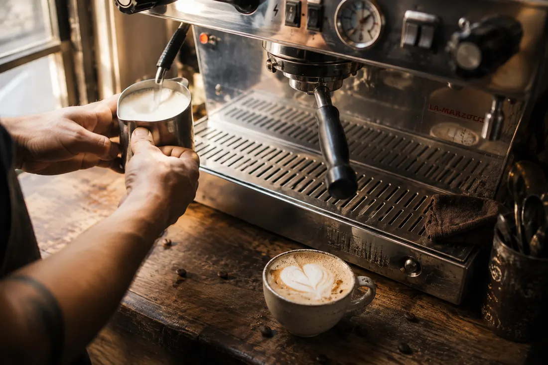 Barista texturando leche junto a la máquina de espresso y una taza con arte latte